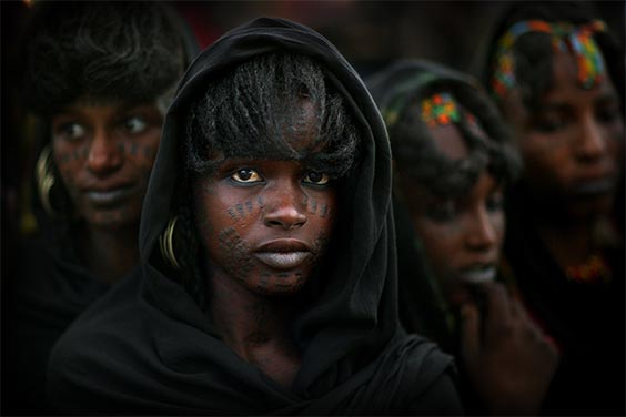 Near Lake Chad, Niger
Among the Wodaabe, pastoral nomads of the sub-Sahara of Niger, charm and beauty are the most desirable character traits of both men and women. Here four women with traditional facial tattoos watch men dancing at the annual Geerewol festival.